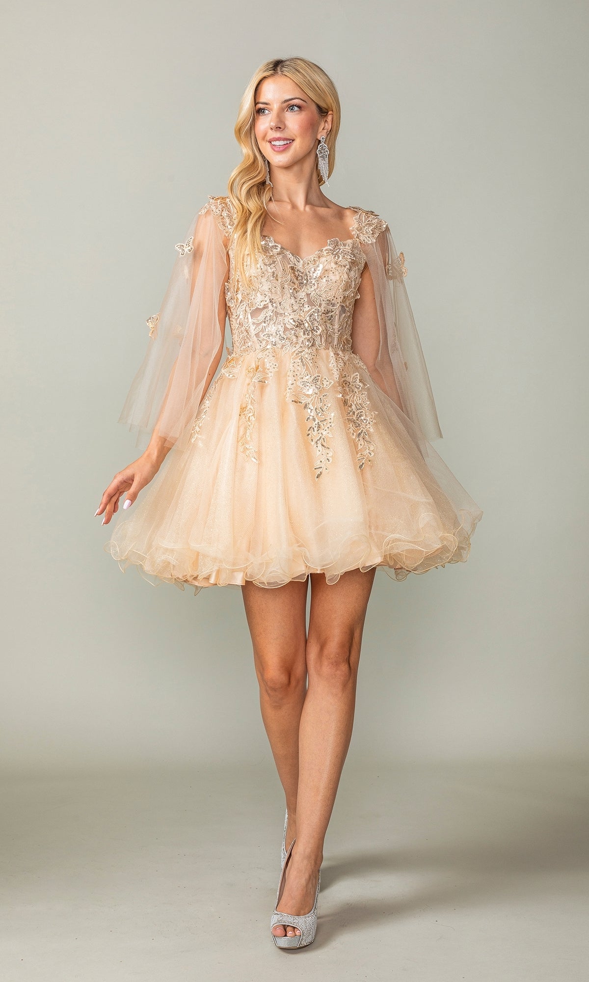 A woman wears the Butterfly-Cape Short A-Line Homecoming Dress 3393 in peach tulle with floral embroidery, sheer sleeves, and sequin appliques, posing against a plain background with a subtle smile.